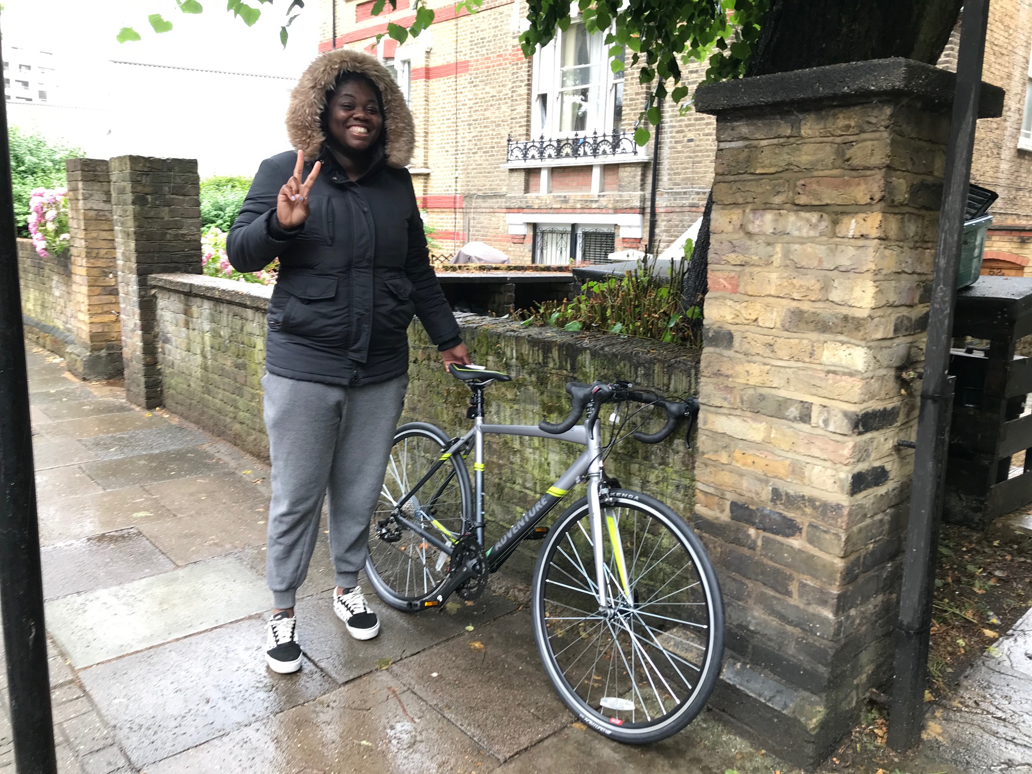 A woman stands on the pavement of a city street with a new road bike alongside, smiling at the camera and flashing a peace sign